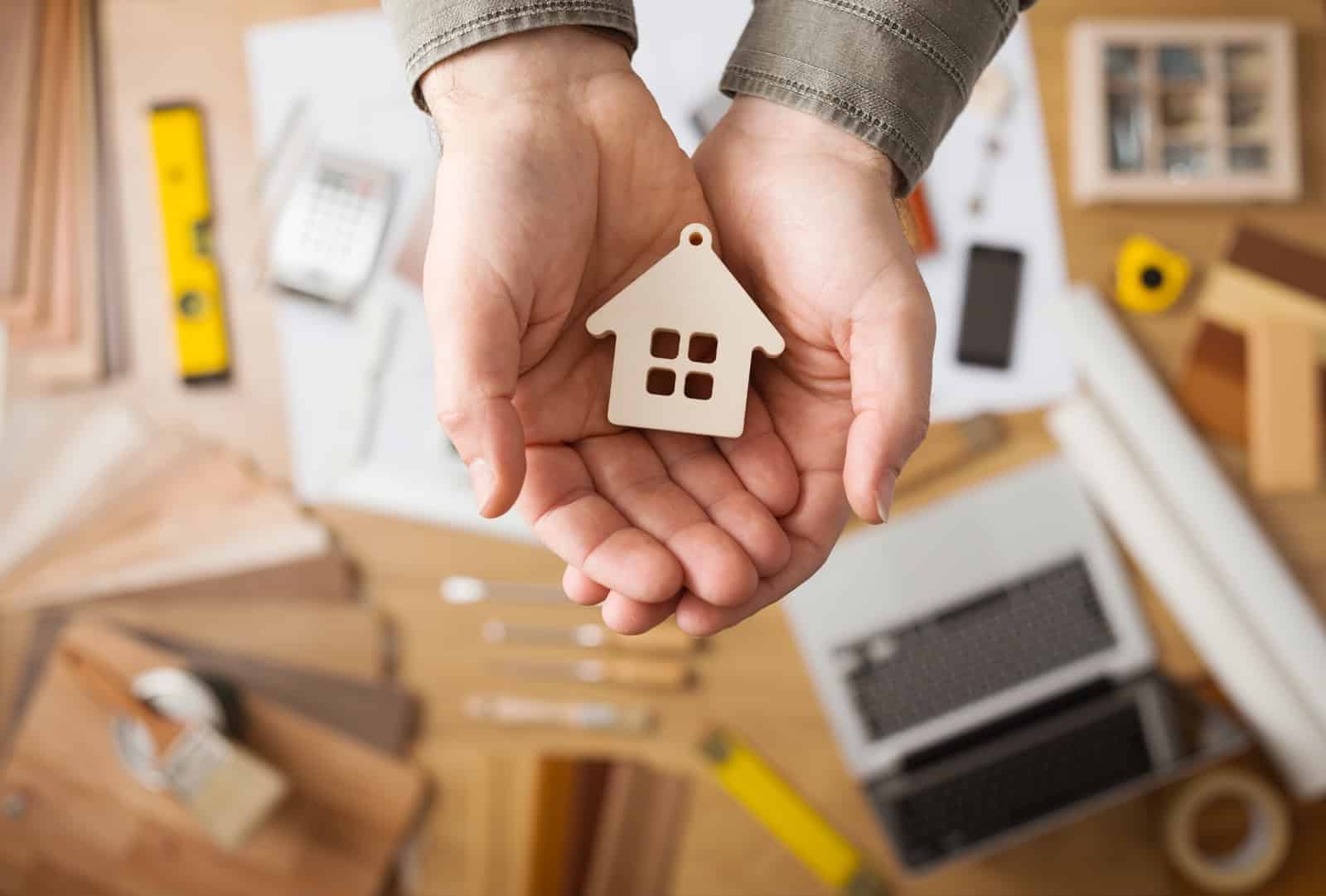 Real estate agent holding a small house, desktop with tools, wood swatches and computer on background, top view